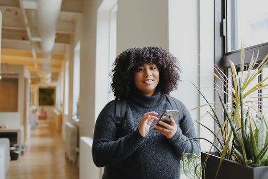 The Hidden Link Between Neurodivergent Burnout and Procrastination—And How to Break It’ a photo of a smiling Black person holding their phone standing next to soothing plants.